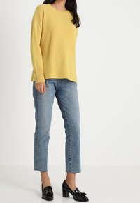 Žlutý texturovaný svetr s kulatým výstřihom, volným střihem a žebrovaným lemem, kombinovaný se světle modrými rovnými džíny a černými botami na blokovém podpatku.
