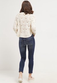 Krämf ärgad stickad tröja med spetsmönster, i kombination med mörkblå åtsittande jeans och ljusbeiga slipskor.