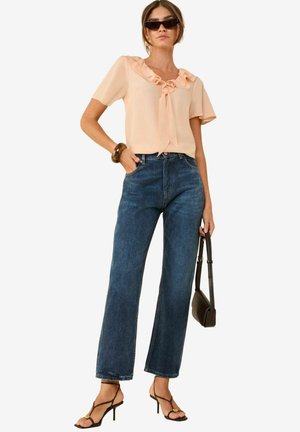 Blouse pêche avec encolure froncée et détail de nœud, associée à un jean en denim bleu taille haute et large, et des escarpins noirs à brides.