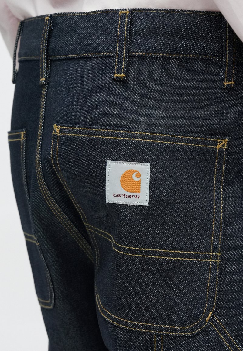 Mörka jeans i denim med framträdande gul sömnad, med bakfickor och en Carhartt-logotyp i vitt och orange på höger bakficka.
