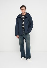 Μπλε αδιάβροχο μπουφάν με κουκούλα, με φερμουάρ μπροστά, φορεμένο πάνω από ένα ριγέ πουλόβερ, συνδυασμένο με φαρδιά τζιν και λευκά αθλητικά παπούτσια.