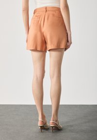 Peach-colored, high-waisted shorts with a smooth texture, cuffed hem, and two back pockets. Paired with beige heeled sandals.