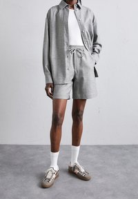 Camicia grigia con bottoni sopra una canotta bianca, abbinata a pantaloni corti grigi. L'outfit presenta un taglio rilassato e un tessuto testurizzato, con sneakers a fantasia.