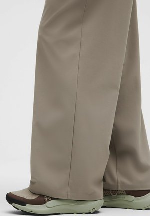 Pantaloni bej cu picior larg, care acoperă gleznele, asortați cu pantofi sport slip-on bej și maro, pe un fundal alb.