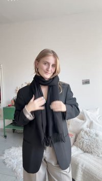 Bufanda negra envuelta alrededor del cuello, combinada con un blazer negro y pantalones de color claro. Textura suave con extremos deshilachados.