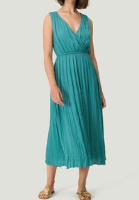 Tyrkis, ærmeløs maxikjole med v-hals og elastisk talje, der har en plisseret tekstur og flydende silhuet. Båret med gyldne sandaler.