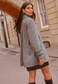 Juodas ir baltas houndstooth modelio paltas su laisvu siluetu, turintis apykaklę, ilgomis rankovėmis ir šoninėmis kišenėmis. Dėvimas virš juodos suknelės ir iki kelių siekiančių botų.