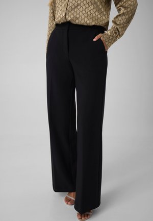 Femme portant un pantalon noir à jambes larges, un chemisier beige à motifs et des talons ouverts à brides, debout avec une main dans la poche.