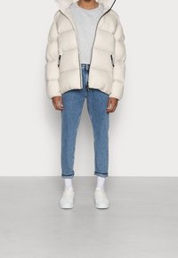 Giacca a vento imbottita di un beige chiaro con finitura lucida, t-shirt grigia sotto, jeans blu e sneakers bianche. Presenta tasche con zip e cappuccio.