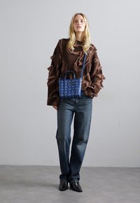 Femme aux longs cheveux blonds portant un pull texturé marron, un jean bleu, des chaussures noires et un sac bandoulière à motifs bleus, se tenant devant un mur gris.