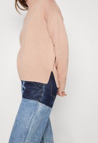 Sweter robiony na drutach w delikatnym różu z wysokim kołnierzem, ściągaczami przy mankietach i bocznymi rozcięciami, zestawiony z jeansami w patchworkowym stylu z jasnoniebieskimi i ciemnoniebieskimi sekcjami.