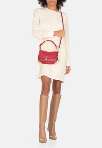 Borsa a tracolla in pelle rossa con forma curvata, hardware dorato e tracolla. Modella che indossa un lungo abito color crema e stivali al ginocchio color beige.
