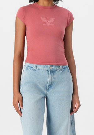 Kvinde iført en tætsiddende lyserød kortærmet top med Guess Originals-logo og lyseblå højtskårne jeans mod en ensfarvet baggrund.