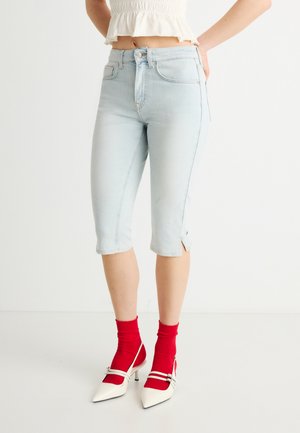 Donna che indossa jeans azzurri scoloriti con spacchi laterali, calzini rossi, tacchi bianchi a punta e un top bianco corto.