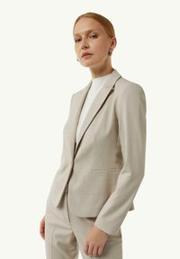 Beżowa marynarka uszyta z teksturowanej tkaniny, z dopasowanym krojem, klapami w kształcie litery V oraz dwoma dyskretnymi kieszeniami, połączona z białą bluzką.