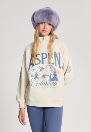 Kremowy polarowy sweter z niebieską grafiką gór i tekstem. Połączony z prążkowanymi niebieskimi legginsami. Fioletowy futrzany kapelusz dopełnia strój.