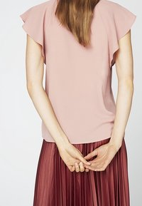Persona vista da dietro che indossa una blusa rosa a maniche corte e una gonna plissettata bordeaux, con le mani unite dietro.
