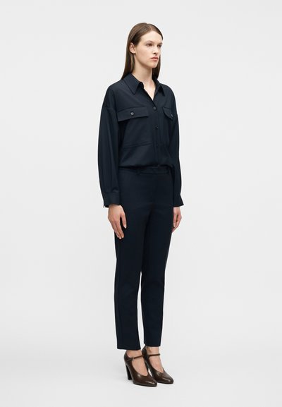 Γυναίκα στέκεται φορώντας σκούρο μπλε πουκάμισο με κουμπιά και τσέπες στο στήθος, εφαρμοστή παντελόνα στο ίδιο χρώμα και σκούρα καφέ παπούτσια Mary Jane με τακούνι.