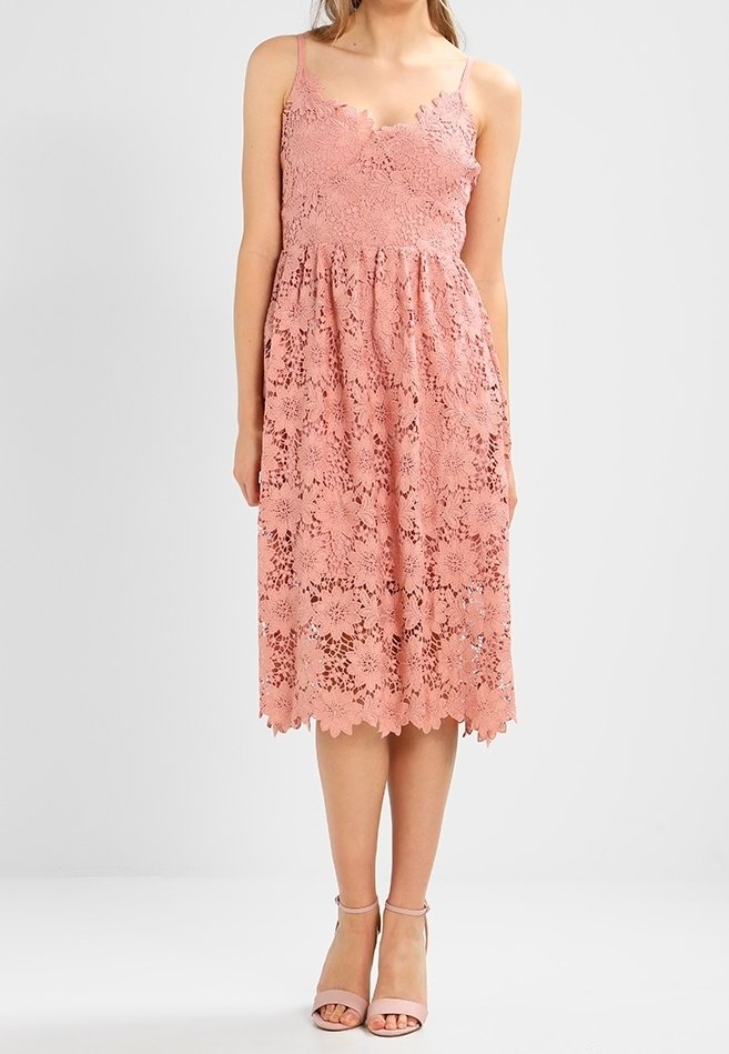 Robe en dentelle rose avec un corsage ajusté, un décolleté festonné et une jupe évasée. Présente des motifs floraux et un ourlet avec des bords festonnés.