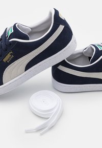 Námornícke semišové tenisky s off-white pruhmi, bielymi gumovými podrážkami a zlatým brandingom; obsahuje bielu šnúrku položenú vedľa topánok.
