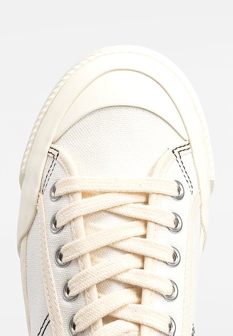 Sneaker bianco in tela con punta in gomma, lacci rotondi e occhielli argentati. Presenta cuciture nere a contrasto lungo i lati.