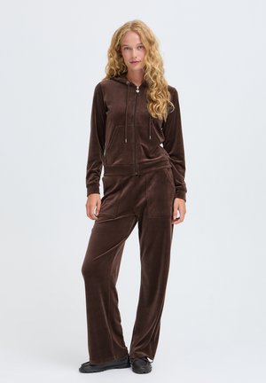 Femme aux longs cheveux bouclés portant un sweat à capuche zippé en velours marron et un pantalon large assorti, debout sur un fond clair uni.