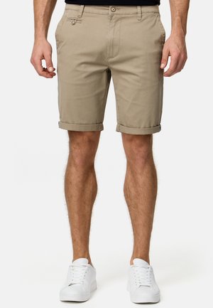 Homme portant un short beige à ourlet retroussé, un haut noir et des baskets blanches, debout devant un fond clair uni.