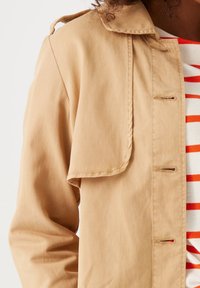 Kurtka w jasnym odcieniu beżu z zapięciem na guziki, dużym kołnierzem i teksturowanym materiałem. Pod spodem widoczna jest biała koszula w czerwone paski.