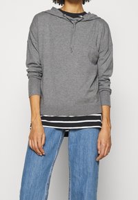 Sweatshirt à capuche gris avec cordon de serrage, ourlet et poignets côtelés, superposé sur un haut rayé noir et blanc, associé à un jean large bleu.