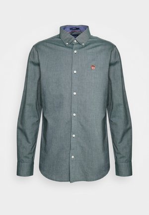 Chemise grise à manches longues boutonnée avec col boutonné et petit logo brodé en forme de bouclier rouge et blanc sur la poitrine gauche.