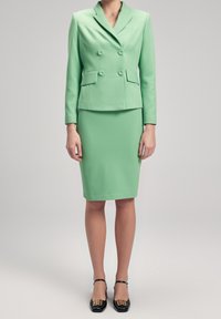 Kvinna iklädd en mintgrön dubbelknäppt kavaj och matchande knälång kjol med svarta klackskor med gulddetaljer.