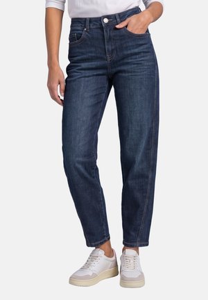 Personne portant un jean taille haute bleu foncé avec des poches avant et des baskets blanches, debout devant un fond uni.