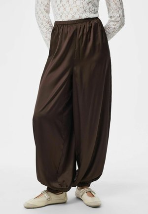 Løse mørkebrune bukser med elastiske mansjetter, brukt sammen med lyse beige Mary Jane-flats og en hvit blondegenser med lange ermer synlig fra livet og nedover.