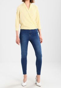 Yellow and white striped wrap top with 3/4 sleeves, paired with dark blue skinny jeans and white high-heeled shoes.