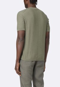 T-shirt verde oliva a maniche corte con collo rotondo a coste, tessuto texturizzato e dettaglio della cucitura centrale; abbinato a pantaloni grigi chiari.