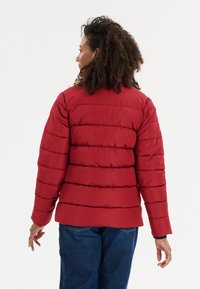 Doudoune rouge avec un design matelassé horizontal, montrant une texture lisse et des poignets élastiques. Portée sur un jean bleu, vue de l'arrière.
