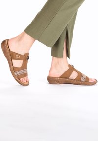 Brown suede sandals with cutouts, adjustable strap, and decorative stitching. Comfortable sole with textured details, paired with olive green pants.