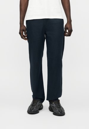 Homme portant un pantalon droit bleu marine et des chaussures de sport noires, debout devant un fond clair uni.