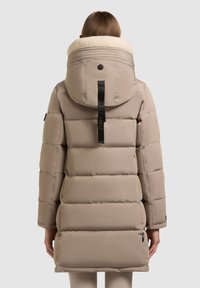 Person, die eine beige gepolsterte Winterjacke mit großer Kapuze trägt, die mit cremefarbenem Fleece gefüttert ist, von hinten vor einem einfarbigen Hintergrund gesehen.