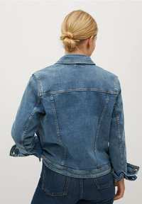 Chaqueta de denim en un azul de lavado medio, con un cuello clásico, puños de botón y un silueta ajustada con detalles de costura en la parte posterior.