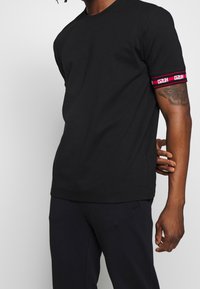 Camiseta de algodón negra con forma ajustada y mangas cortas. Presenta detalles a rayas rojas y blancas en la manga. Diseño minimalista.