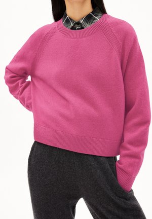 Femme portant un pull rose vif par-dessus une chemise à carreaux noir et blanc, associée à un pantalon gris foncé, posant avec une main sur la hanche.