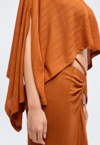 Bras et torse latéral portant un haut en tricot côtelé orange brûlé et une jupe avec des détails froncés et des découpes, sur un fond clair uni.