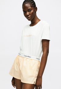 Γυναίκα που φοράει λευκό t-shirt Calvin Klein και ροδακινί σορτς με ελαστική μέση Calvin Klein, στέκεται μπροστά σε απλό φωτεινό φόντο.