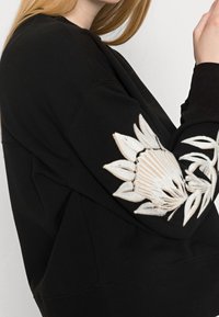 Czarna bluza z intricate, jasnym haftem kwiatowym na lewym rękawie, w luźnym kroju i miękkiej teksturze.