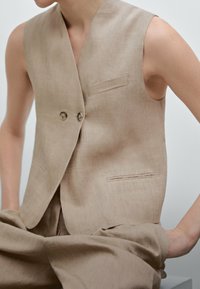 Beige linneställ med djup V-ringning, två knappar och sidofickor. Slät textur och minimalistisk design, i kombination med matchande byxor.