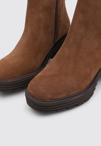 Bottines en daim marron avec un bout arrondi, fermeture éclair sur le côté et semelle épaisse à motifs. Texture lisse avec une finition mate.