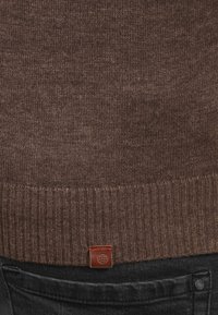 Brun strikket genser med ribbet kant. Har et lite lærmerke nederst. Mørke jeans synlig under genseren.