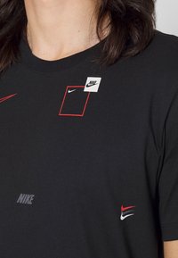 Camiseta de algodón negra con varios logotipos de Nike en rojo y blanco, incluyendo un parche cuadrado y diferentes estilos del característico swoosh de la marca.