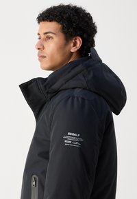 Chaqueta aislante negra con un cuello alto, capucha y cierre frontal con cremallera. Presenta una marca visible y elementos de diseño funcionales en la manga.